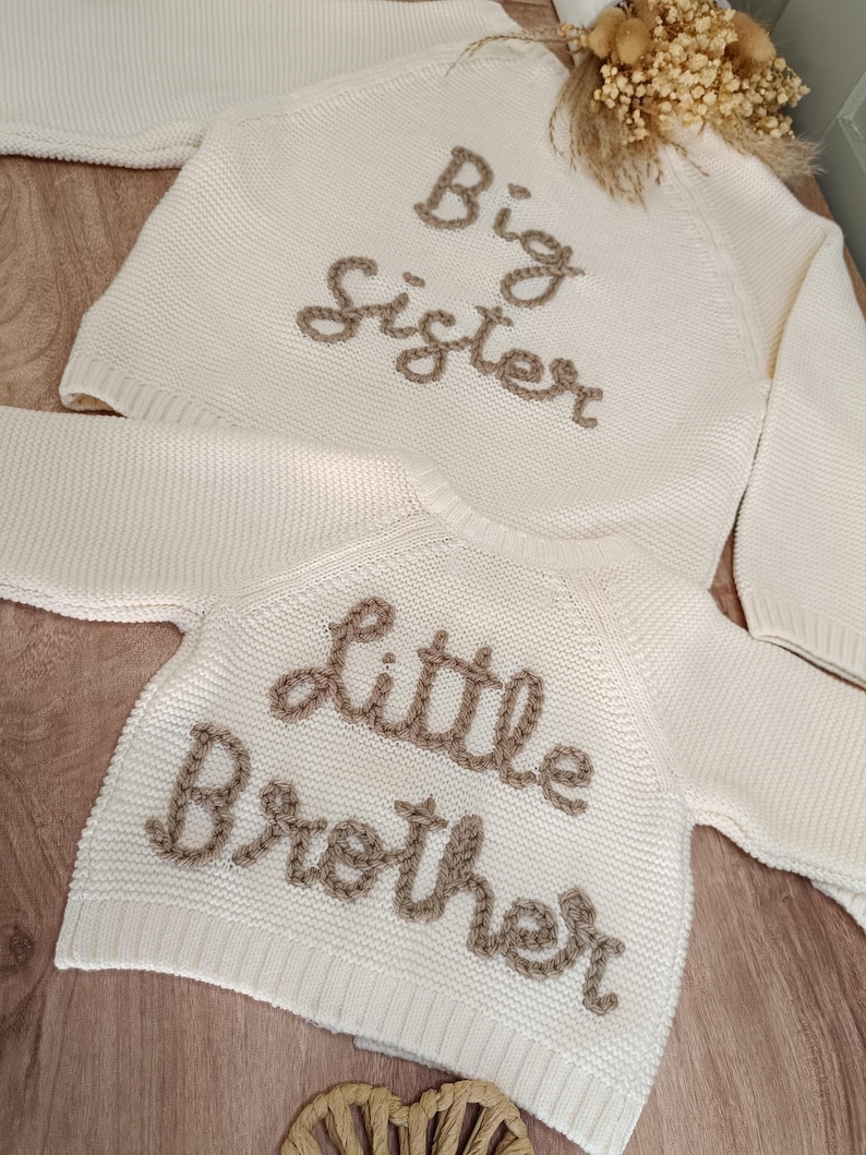 K&ouml;nnte beinhalten: Cremefarbene Strickpullover, einer mit der Aufschrift "Big Sister" und der andere mit "Little Brother" in brauner Stickerei. Die Pullover liegen auf einer hellen Holzoberfl&auml;che, mit einem kleinen Blumengesteck im Hintergrund.