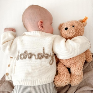 May include: A baby wearing a cream-colored knitted cardigan with the word "Saturday" embroidered on the back, paired with striped pants, cuddles a brown teddy bear. The bear has a yellow tag.