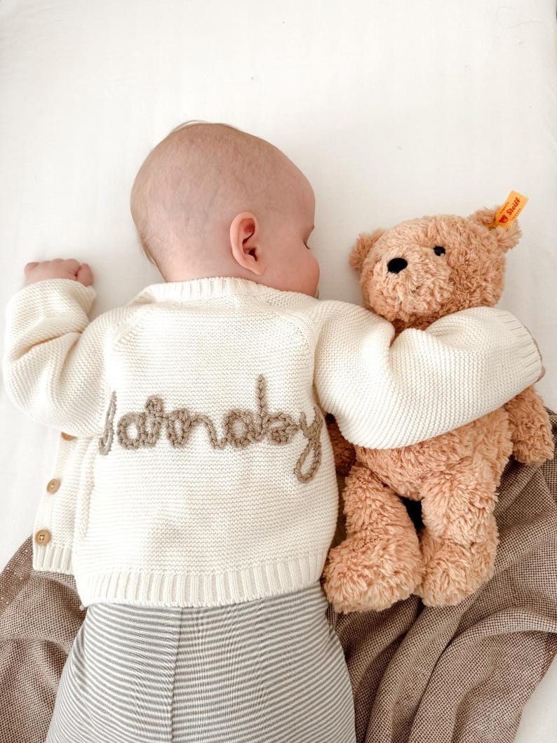 May include: A baby wearing a cream-colored knitted cardigan with the word "Saturday" embroidered on the back, paired with striped pants, cuddles a brown teddy bear. The bear has a yellow tag.