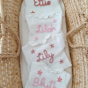 May include: Four white knitted baby blankets with names embroidered in pink: "Ettie", "Lilah", "Lily", and "Blythe". Each blanket has a ruffled edge and pink flower accents. The blankets are arranged in a woven basket.