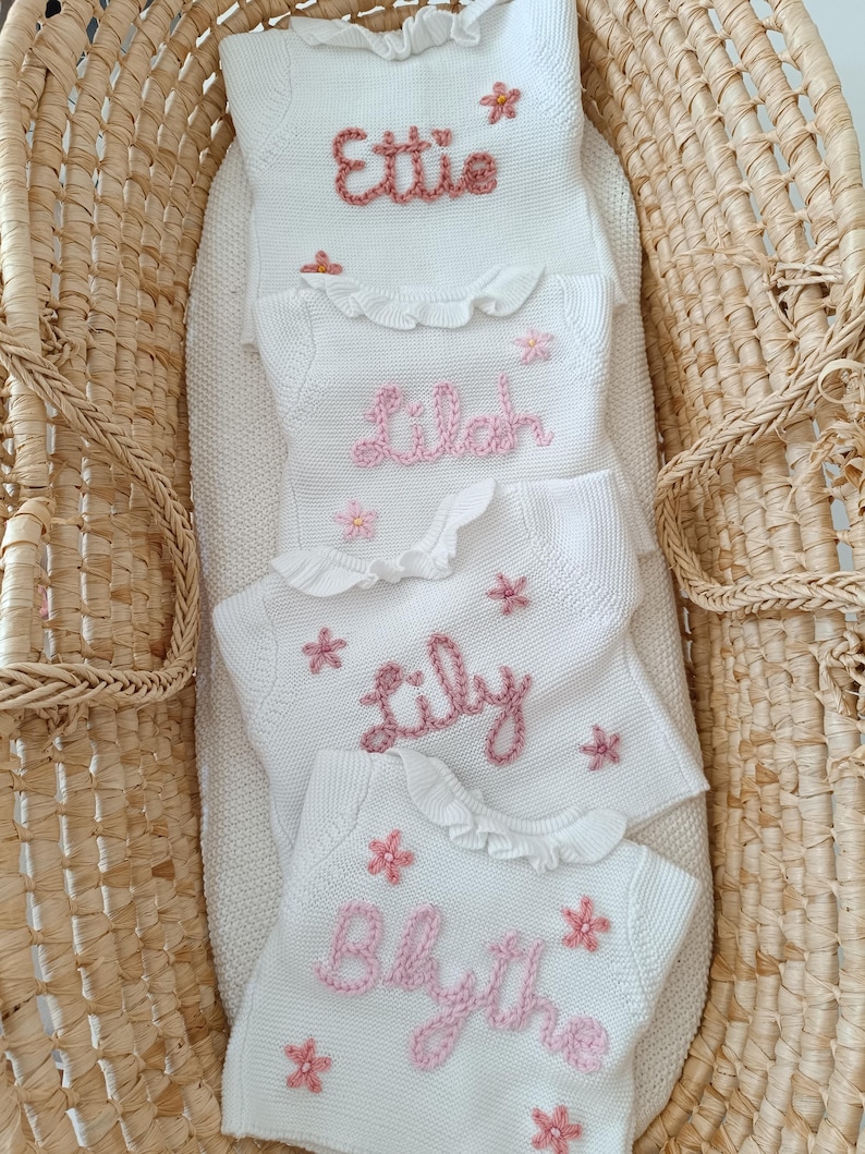 May include: Four white knitted baby blankets with names embroidered in pink: "Ettie", "Lilah", "Lily", and "Blythe". Each blanket has a ruffled edge and pink flower accents. The blankets are arranged in a woven basket.