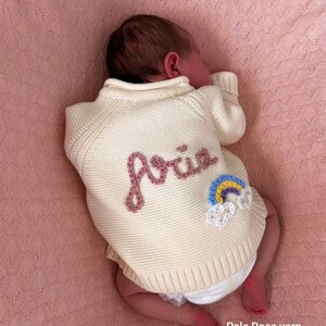 K&ouml;nnte beinhalten: Ein cremefarbener Baby-Pullover mit dem Namen "Aria" in Rosa gestickt. Ein kleiner Regenbogen-Aufn&auml;her ist auf dem R&uuml;cken aufgen&auml;ht. Das Baby liegt auf einer rosa Decke. Der Text "Pale Rose yarn Small add on" ist sichtbar.