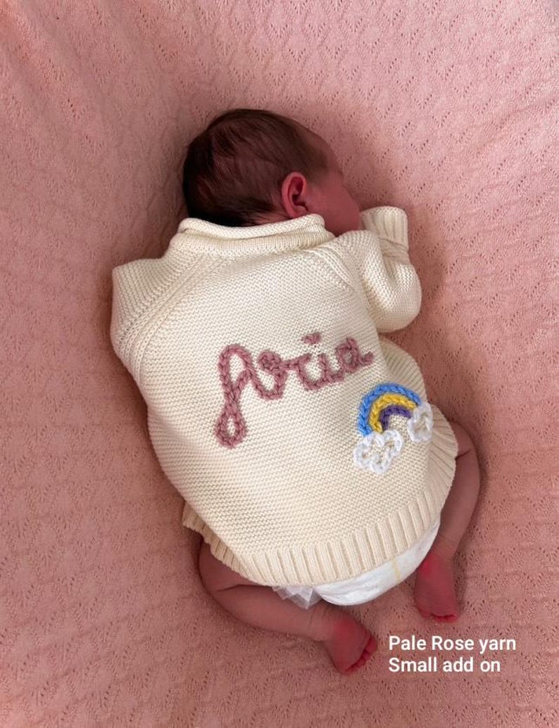 K&ouml;nnte beinhalten: Ein cremefarbener Baby-Pullover mit dem Namen "Aria" in Rosa gestickt. Ein kleiner Regenbogen-Aufn&auml;her ist auf dem R&uuml;cken aufgen&auml;ht. Das Baby liegt auf einer rosa Decke. Der Text "Pale Rose yarn Small add on" ist sichtbar.