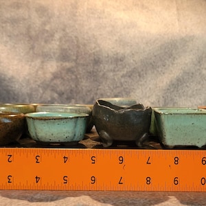 May include: A collection of small ceramic pots in various shapes and colors, including brown, green, and gray. The pots are arranged on a black surface, with a ruler showing measurements in inches. These miniature pots are ideal for succulents or small plants.