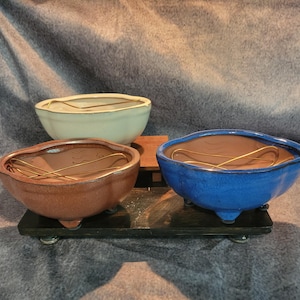 May include: Three ceramic bonsai pots in a row. The pots are in shades of brown, blue, and off-white. Each pot has a unique shape and a wire structure inside. The pots are on a black wooden stand.