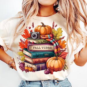 Könnte beinhalten: Cremefarbenes T-Shirt mit einem Herbst-Themen-Grafik. Das Design zeigt einen Stapel Bücher, Kürbisse, Herbstblätter und eine Kamera. Die Worte "FALL VIBES" sind auf den Büchern aufgedruckt. Das Shirt ist eine PNG-Datei für den kommerziellen Gebrauch.