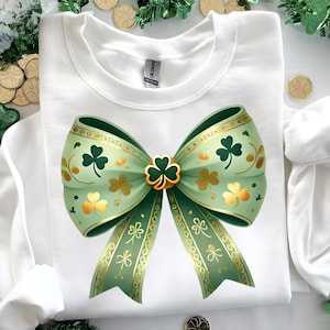 May include: White sweatshirt featuring a large green bow with gold accents and shamrock designs. The bow has a central shamrock clasp and decorative gold trim. A festive St. Patrick's Day themed design.