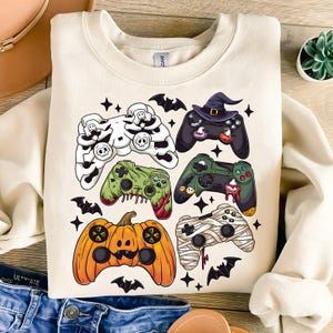 May include: Cream-colored sweatshirt featuring a Halloween-themed design. The design includes various game controllers decorated as a skull, zombie, witch, mummy, and pumpkin. Black bats and stars are scattered around the controllers.