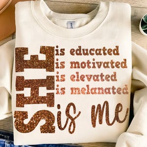 May include: Cream-coloured sweatshirt with the words "is educated, is motivated, is elevated, is melanated, is me" in a glittery brown font. The design features a puzzle piece graphic.