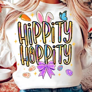 Może przedstawiać: Biała bluza z napisem "HIPPITY HOPPITY" w kolorowych literach. Projekt zawiera marchewkę, uszy królika, motyla, ślady łap, fioletową kokardę i pisanki. Idealna na Wielkanoc.
