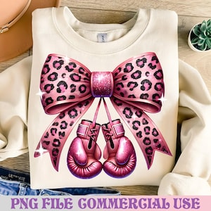 以下が含まれることがあります： クリーム色のスウェットシャツ。ヒョウ柄のピンク色の大きなリボンとキラキラしたアクセントが特徴です。リボンの下には、2つのピンク色のボクシンググローブがあります。「PNG FILE COMMERCIAL USE」というテキストが下にあります。