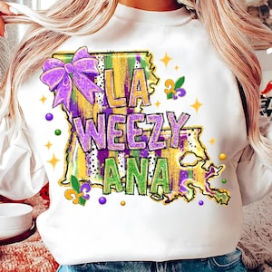May include: White sweatshirt featuring a purple bow and the text "LA WEEZY ANA" in purple and green. The design includes a map outline of Louisiana with yellow, green, and purple stripes, and star and fleur-de-lis accents.