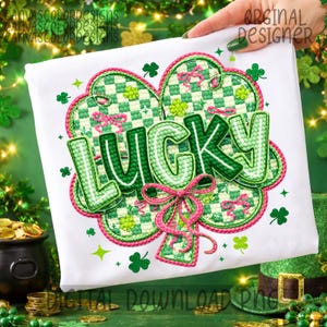 Può includere: T-shirt bianco con un design ricamato verde e rosa. Il design include un quadrifoglio, la parola "LUCKY" in verde e un fiocco rosa. La maglietta è decorata con trifogli e stelle, perfetta per San Patrizio.