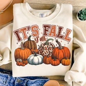 May include: Cream-colored sweatshirt with the text "IT'S FALL y'all" in brown and orange. The design features various pumpkins in shades of orange, white, and brown, some with leopard print patterns. A seasonal autumn-themed graphic.