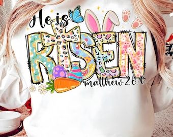 May include: White sweatshirt with a colorful Easter-themed graphic. The design includes the words "He is Risen" in a mix of fonts and patterns, a cross, bunny ears, eggs, a carrot, and a butterfly. The text "matthew 28:6" is also included.
