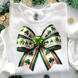 May include: White sweatshirt featuring a large, decorative bow with green and white ribbons. The bow is adorned with shamrocks and gold accents, with green and orange stripes. A large shamrock is at the center.