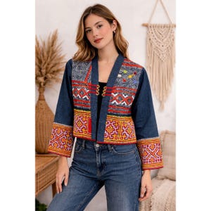 Peut inclure: Veste en jean bleue avec des détails brodés colorés. La veste présente des motifs géométriques rouges, jaunes et blancs sur les manches et les panneaux avant. Elle a un design sans col et trois fermetures décoratives.