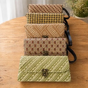 Puede incluir: Cinco bolsos de mano rectangulares tejidos en tonos marrón, amarillo y verde están apilados sobre una superficie de madera. Cada bolso tiene un cierre de broche y una correa negra. Los bolsos tienen diferentes patrones tejidos.