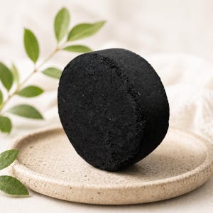 May include: A solid, circular black cleansing bar with a textured, porous surface. The round, dark disc is positioned upright on a small, speckled beige ceramic plate. The object has a uniform, matte appearance and a thick, cylindrical shape.