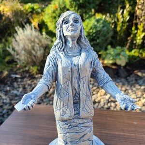 Puede incluir: Una estatua de cerámica azul de una mujer con el pelo largo, de pie con los brazos extendidos. La estatua está detallada con una superficie texturizada, que se asemeja a un patrón de remolino.