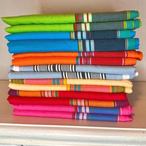 May include: A stack of colorful, folded textiles. The textiles are in various colors, including green, blue, orange, red, pink, and yellow. Each textile has a striped pattern in the center with a variety of colors. The textiles are stacked on a white surface.