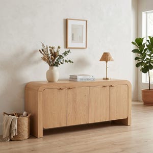 Puede incluir: Un mueble de madera de roble claro con cuatro puertas, frente curvo y acabado de color claro. Una pequeña lámpara tejida, un jarrón con flores secas y libros están encima. También se ven una cesta tejida y una impresión enmarcada.