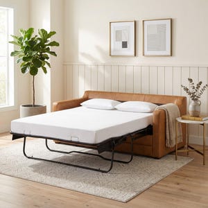 May include: A brown leather sofa bed with a white mattress is extended. Two white pillows rest on the mattress. A beige throw blanket is draped over the arm. A small side table with a marble top and gold legs is next to the sofa.