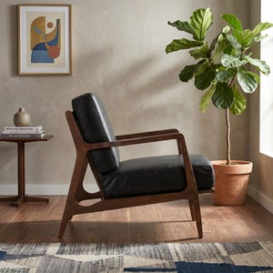 Puede incluir: Un sillón moderno con asiento de cuero negro y estructura de madera oscura. La silla tiene un diseño minimalista con reposabrazos y patas en ángulo. Una planta en maceta y una obra de arte se encuentran en el fondo, creando un ambiente interior elegante.