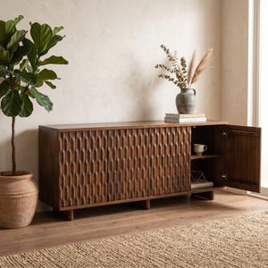 Può includere: Una credenza in legno marrone scuro con un design geometrico intagliato sulle ante. L'armadio ha una forma rettangolare e poggia su gambe corte. Sono visibili una pianta in vaso di terracotta e un vaso con fiori secchi.