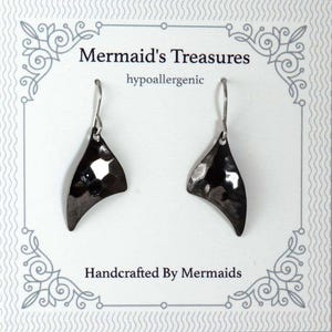 May include: A pair of handcrafted, hypoallergenic, black hammered metal earrings. The earrings are in the shape of a mermaid's tail fin. The card behind the earrings reads "Mermaid's Treasures" and "Handcrafted By Mermaids".