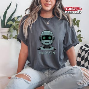 以下が含まれることがあります： 漫画のロボットと「MARVIN」というピクセル化された文字がプリントされた濃いグレーのTシャツ。 ロボットは水色で、黒い目と悲しそうな表情をしています。 シャツの袖はロールアップされています。