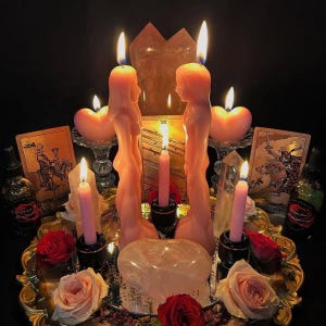 Puede incluir: Dos velas rosas con forma humana se enfrentan sobre una bandeja dorada, rodeadas por cuatro velas cónicas rosas, dos velas en forma de corazón, rosas rojas y rosas, dos cartas del tarot, dos frascos de vidrio y un gran cristal en forma de corazón.