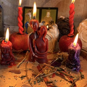 Puede incluir: Una colección de velas encendidas sobre una bandeja plateada, que incluye una figura de cera roja de dos personas abrazadas, dos velas rojas retorcidas en manzanas, una pequeña vela de Buda roja y una vela morada con hierbas secas.