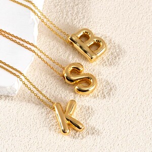 May include: Gold-colored initial necklaces. The necklaces feature the letters B, S, and K. Each letter is a bold, three-dimensional design suspended from a delicate gold chain. The background is a neutral, textured surface.