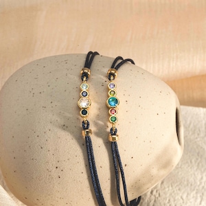 May include: Two adjustable bracelets with black cords and gold accents. Each bracelet features a row of colourful gemstones.