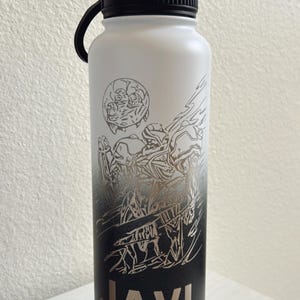 May include: A black and white stainless steel water bottle with a gradient design. The design features a stylized illustration of a creature with a large head and a name "JAVI" printed on the bottom.