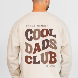 Könnte beinhalten: Beiges Sweatshirt mit dem Text "PROUD MEMBER COOL DADS CLUB EST 2025" in braunen und beigen Buchstaben. Das Sweatshirt hat einen Rundhalsausschnitt und lange Ärmel.