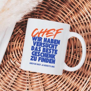Peut inclure: Mug en céramique blanche avec le mot "CHEF" en orange et du texte bleu qui dit "WIR HABEN VERSUCHT DAS BESTE GESCHENK ZU FINDEN ABER DU HAST JA BEREITS UNS". La tasse a une anse incurvée et est posée sur une surface tissée.