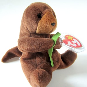 May include: Brown plush otter beanie baby toy holding a green leaf. The toy has a red heart-shaped tag with the words "Ty Inc." and "Beanie Babies" printed on it.