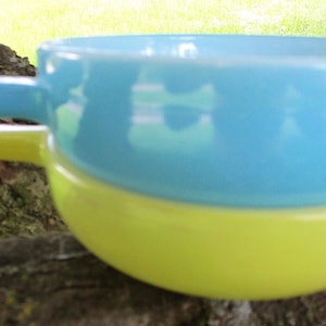 May include: A stack of two ceramic bowls, one light blue and one yellow, each with a handle. The bowls are set against a blurred background of a tree branch and green foliage.