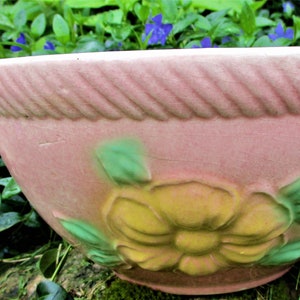 May include: A pink ceramic bowl with a rope design around the rim. The bowl has a floral design with a yellow flower and green leaves.