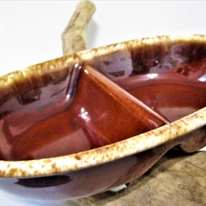 May include: A brown ceramic divided serving dish with a glossy finish. The dish has a unique shape with a central divider and a speckled, light brown rim. The dish is resting on a piece of wood.