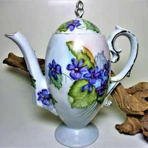 May include: A white porcelain teapot with a delicate floral design featuring purple violets and green leaves. The teapot has a gold trim and a lid with a knob.