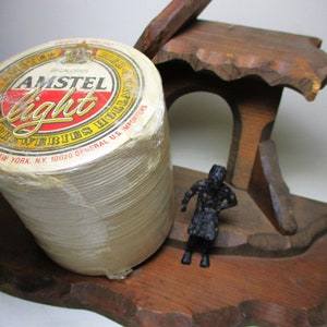 May include: A stack of Amstel Light beer coasters in a wooden holder. The coasters are wrapped in plastic and have the Amstel Light logo printed on them. The holder is shaped like a small house with a figure standing in the doorway.