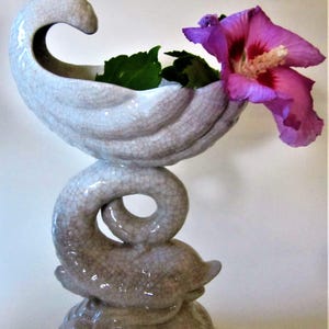 May include: A white ceramic vase shaped like a dolphin with a shell-shaped top. The vase is decorated with a crackled glaze and holds a single pink flower and green leaves.