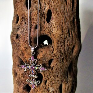 May include: A silver chain necklace with a pink rhinestone cross pendant. The pendant has a detailed design with swirls and a large center stone.
