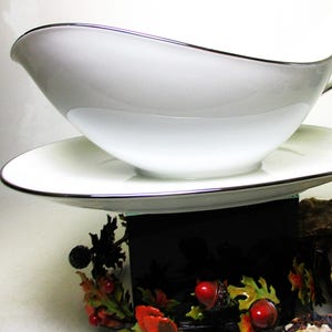 May include: A white gravy boat and saucer set with a silver trim. The gravy boat has a curved handle and a spout. The saucer is round. The set is displayed on a decorative black stand with colorful floral accents.