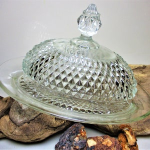 Puede incluir: Un plato de mantequilla de vidrio transparente con un patrón de diamante. El plato tiene una tapa con un pomo en la parte superior. El plato está sentado sobre un trozo de madera a la deriva.