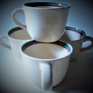 May include: Four white ceramic coffee mugs stacked together. Each mug has a rounded shape with a handle and a decorative rim featuring a teal, burgundy, and dark brown stripe. The mugs are arranged in a pyramid shape.