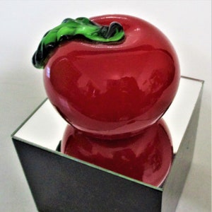 May include: A red glass apple with a green leaf on top, sitting on a black square with a mirrored surface.